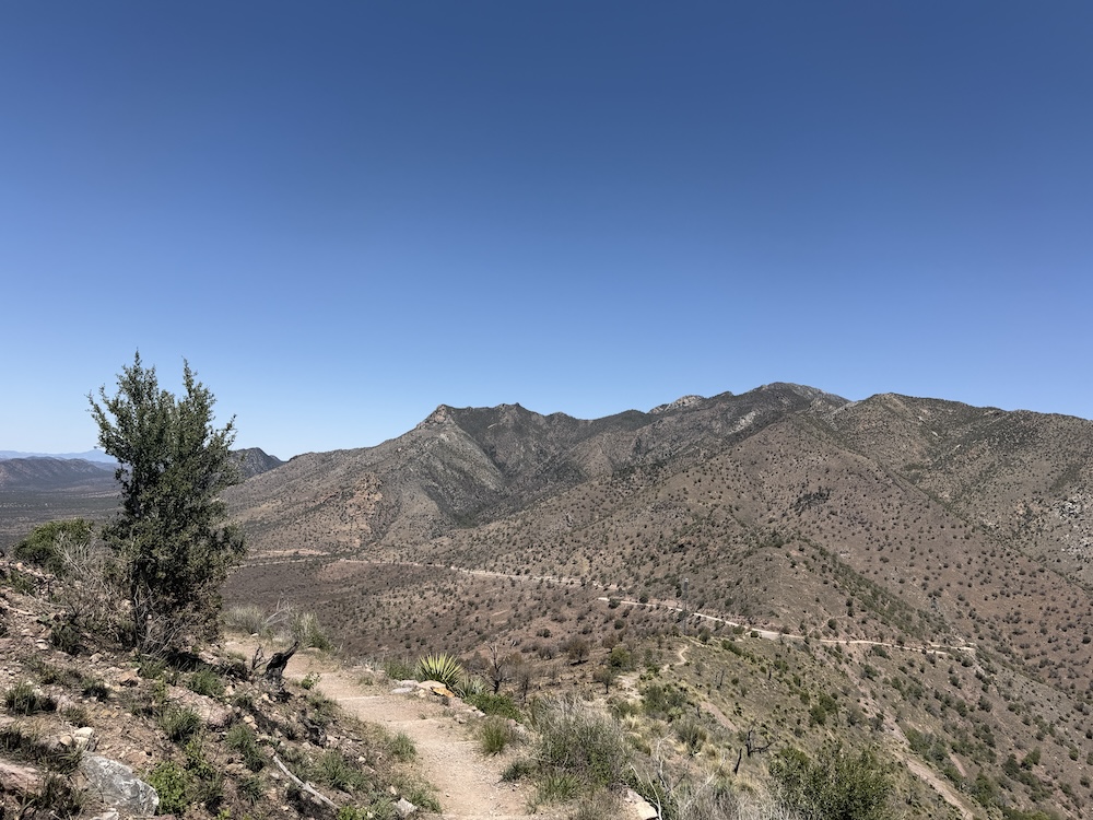 A dirt hiking trail winds through dry, rocky hills with scattered bushes and a clear blue sky above. Mountains stretch across the background.