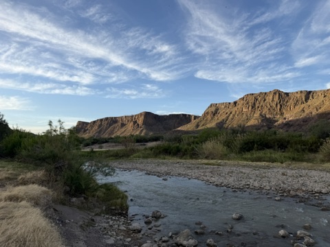 A shallow river flows past rocky banks with brush and grass, set against rugged, sunlit cliffs under a blue sky with wispy clouds.